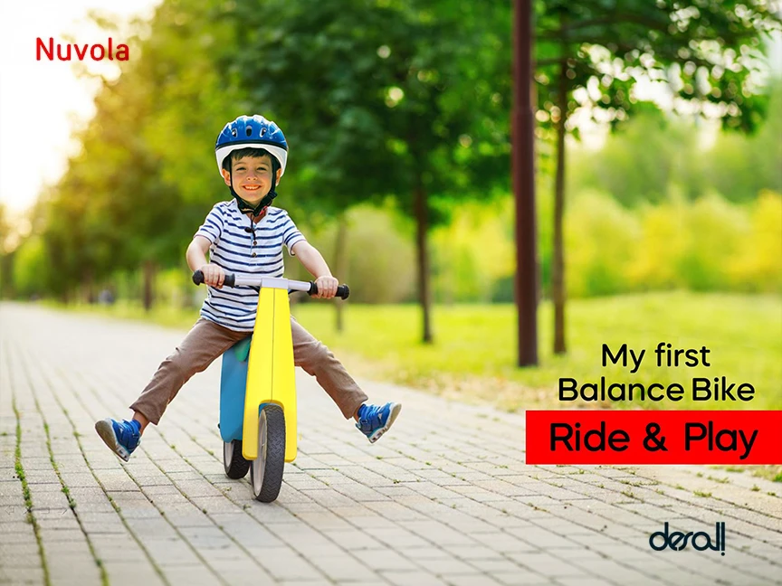 Child in a helmet riding the yellow Nuvola balance bike along a tree-lined park path
